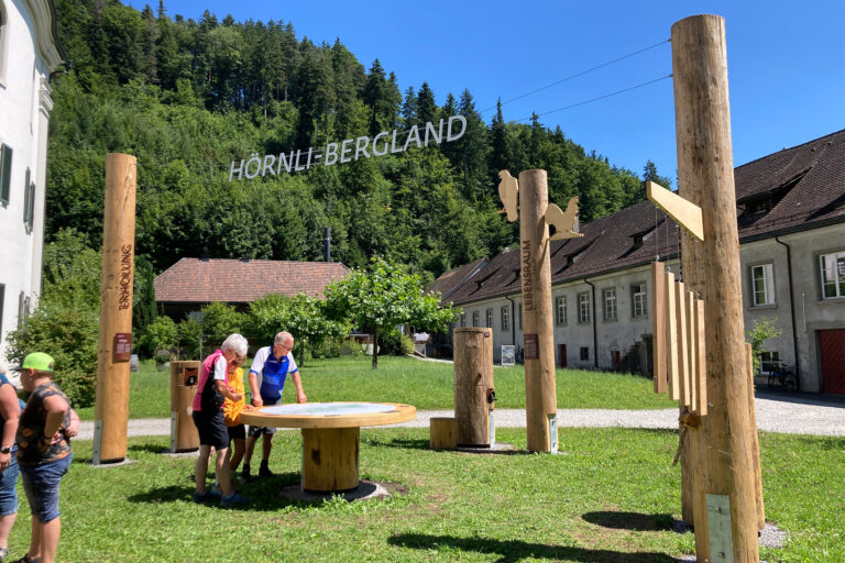 Open meeting place with large wooden steles, interactive stations, and a round information table, surrounded by green landscape and historic monastery buildings.