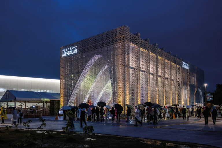 Der Aserbaidschan-Pavillon an der Expo 2025 in Osaka bei Nacht. Das Gebäude ist von einer filigranen, ornamentalen Fassade umhüllt, die von innen stimmungsvoll beleuchtet wird. Besucherinnen und Besucher mit Regenschirmen stehen in einer Schlange vor dem Eingang.